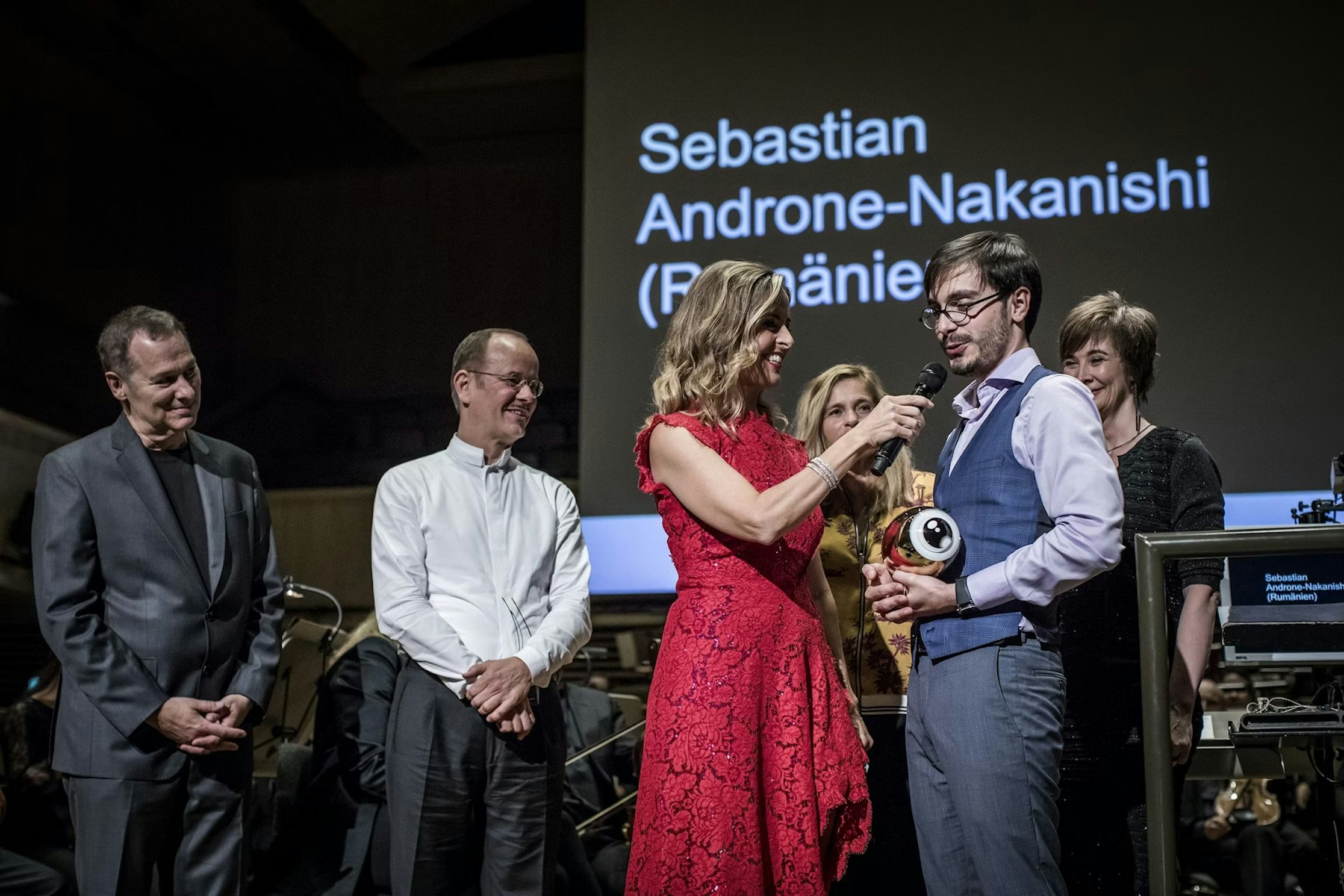 Eine Frau in einem roten Kleid steckt einem lächelnden Mann mit einer Trophäe eine Auszeichnung an, während vier Personen in der Nähe auf der Bühne stehen. Im Hintergrund ist Sebastian Androne-Nakanishi (Rumänien) zu sehen.