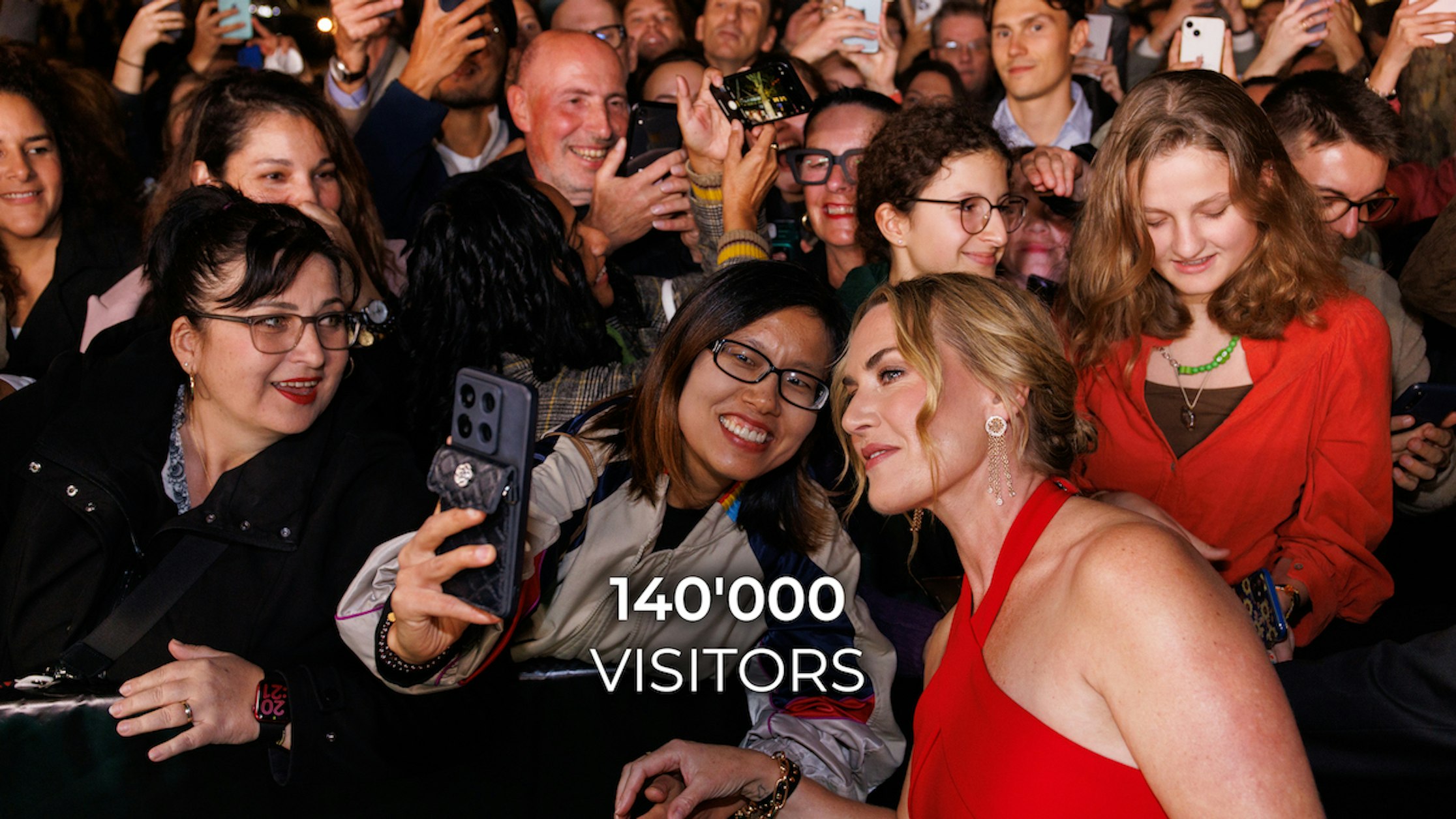 Eine lächelnde Frau in einem roten Kleid macht Selfies mit aufgeregten Fans in einer überfüllten, festlichen Umgebung. Der Text auf dem Bild lautet: 140.000 BESUCHER.