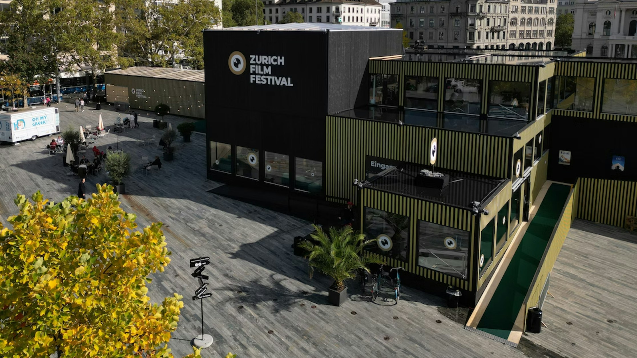 Ein modernes schwarz-grünes Gebäude mit der Aufschrift „Zürich Film Festival“ steht im Tageslicht auf einem weitläufigen Platz mit Menschen, Bäumen und umgebender Stadtarchitektur.