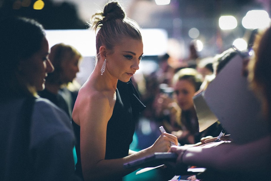 Eine Frau in einem schwarzen Kleid mit einem Dutt gibt bei einer Veranstaltung Autogramme für Fans, umgeben von Menschen und hellen Lichtern im Hintergrund.