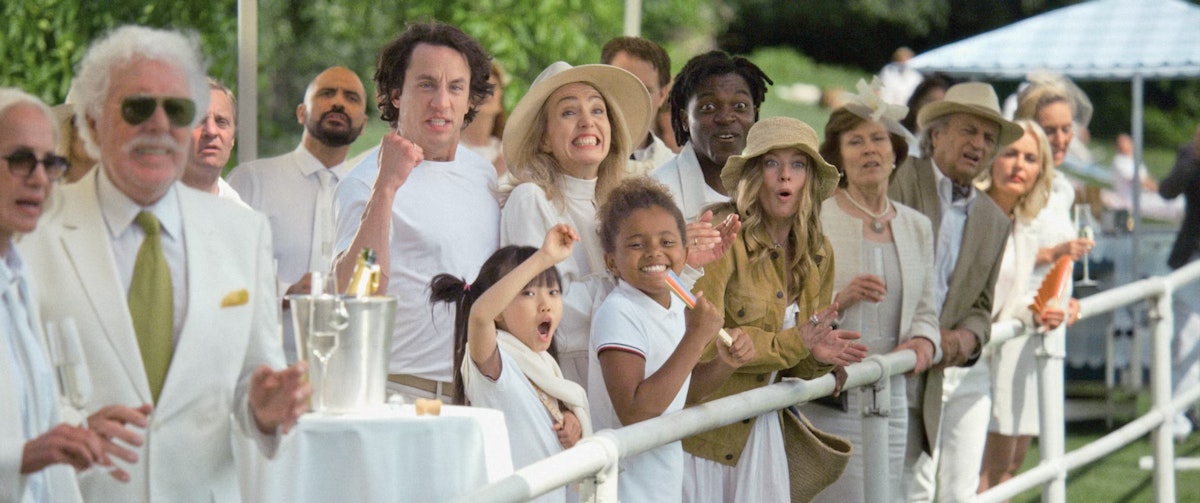 A diverse group of people dressed in white and beige summer clothing stand by a railing outdoors, cheering and watching an event with excitement and surprise. Some raise their arms or fists in celebration.