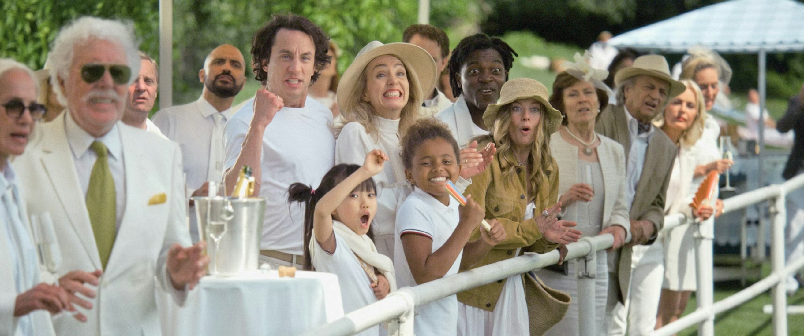 A diverse group of people dressed in white and beige summer clothing stand by a railing outdoors, cheering and watching an event with excitement and surprise. Some raise their arms or fists in celebration.
