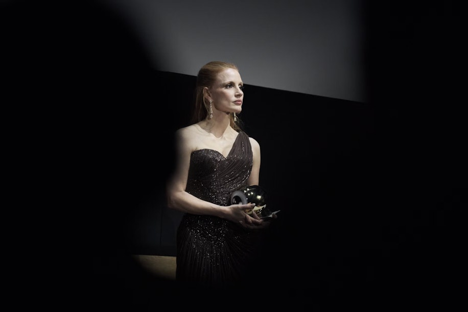 Eine Frau in einem eleganten, dunklen, einschultrigen Kleid steht mit einer glänzenden Auszeichnung in der Hand da und blickt zur Seite. Dramatische Beleuchtung und dunkle Schatten rahmen das Bild ein.