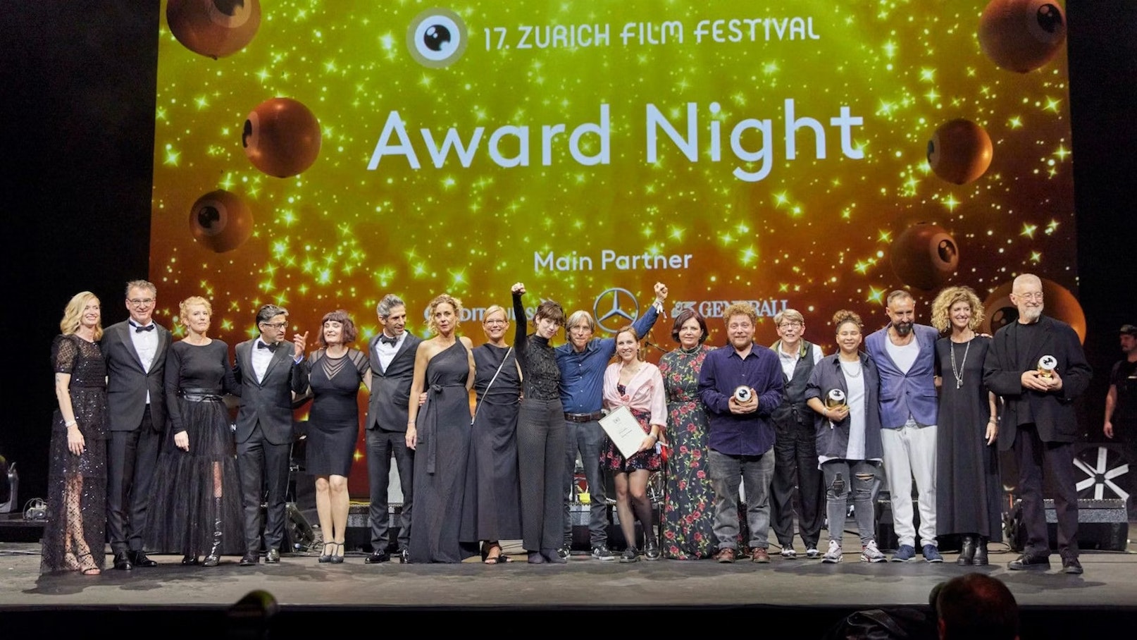 Bei der Award Night des Zurich Film Festival posiert eine große Gruppe von Menschen auf der Bühne. Einige halten Trophäen in den Händen, vor einem glitzernden Goldhintergrund, auf dem der Name der Veranstaltung und die Sponsoren zu sehen sind.