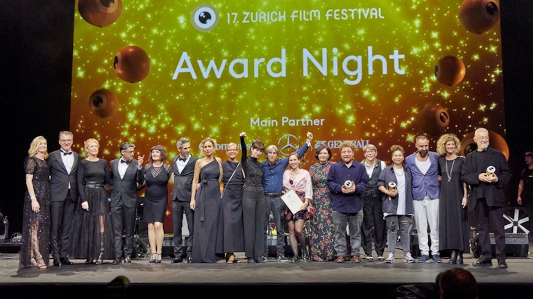 Bei der Award Night des Zurich Film Festival posiert eine große Gruppe von Menschen auf der Bühne. Einige halten Trophäen in den Händen, vor einem glitzernden Goldhintergrund, auf dem der Name der Veranstaltung und die Sponsoren zu sehen sind.