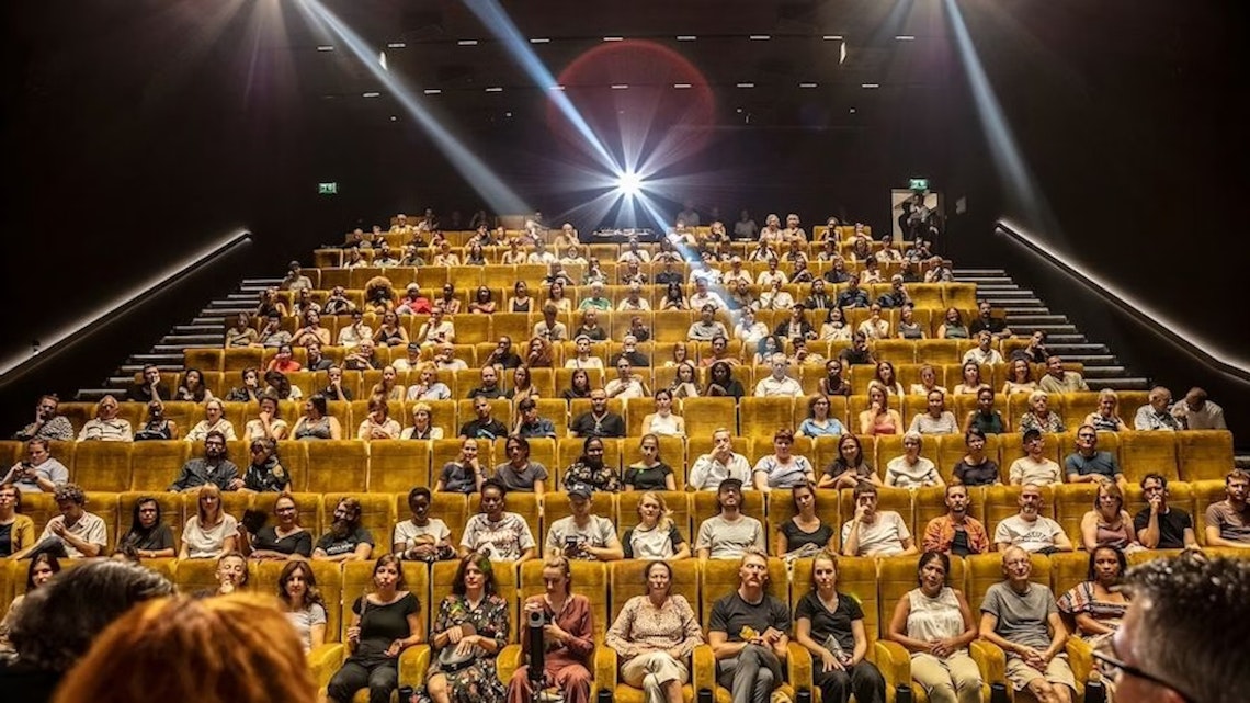 A large audience sits in yellow theater seats, facing the camera in a well-lit auditorium. Bright lights shine from the back, illuminating the crowd and creating a dramatic atmosphere.