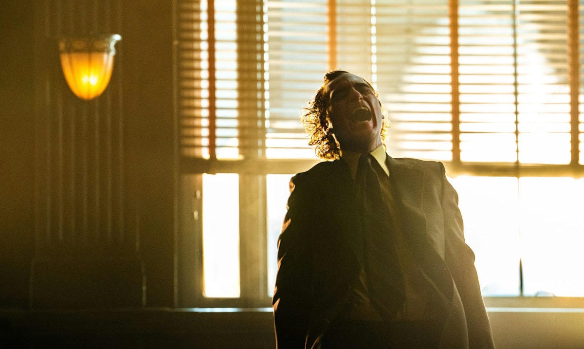 A man in a suit stands in front of a sunlit window with blinds, head tilted back and mouth open as if laughing or shouting, with dramatic lighting casting shadows in the room.