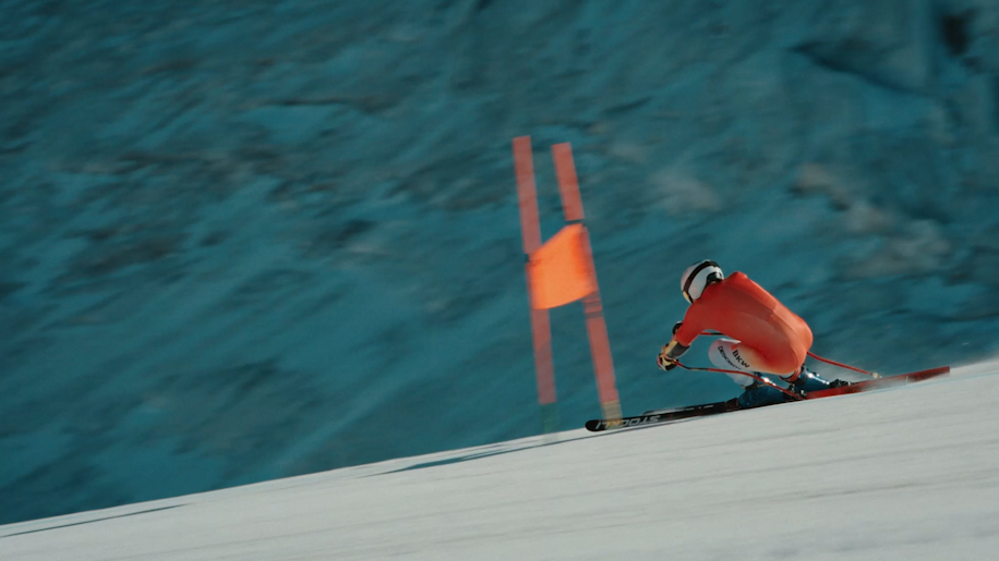 A skier in a red suit and helmet makes a sharp turn around an orange flag on a snowy slope, with a blurred blue-gray mountain background.