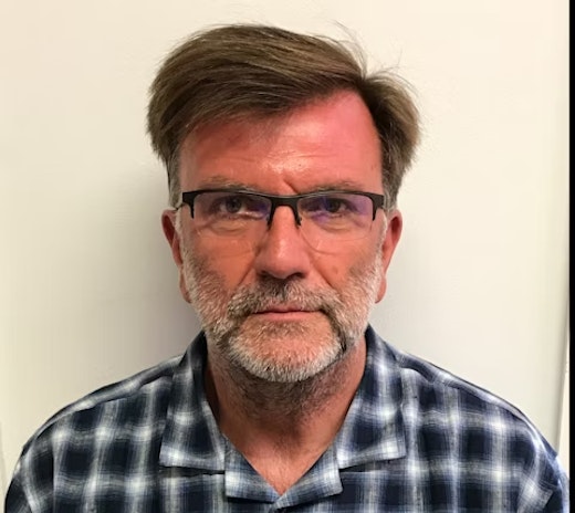 A middle-aged man with short brown hair, glasses, and a gray beard is wearing a blue and white plaid shirt and standing against a plain white background, looking directly at the camera with a neutral expression.