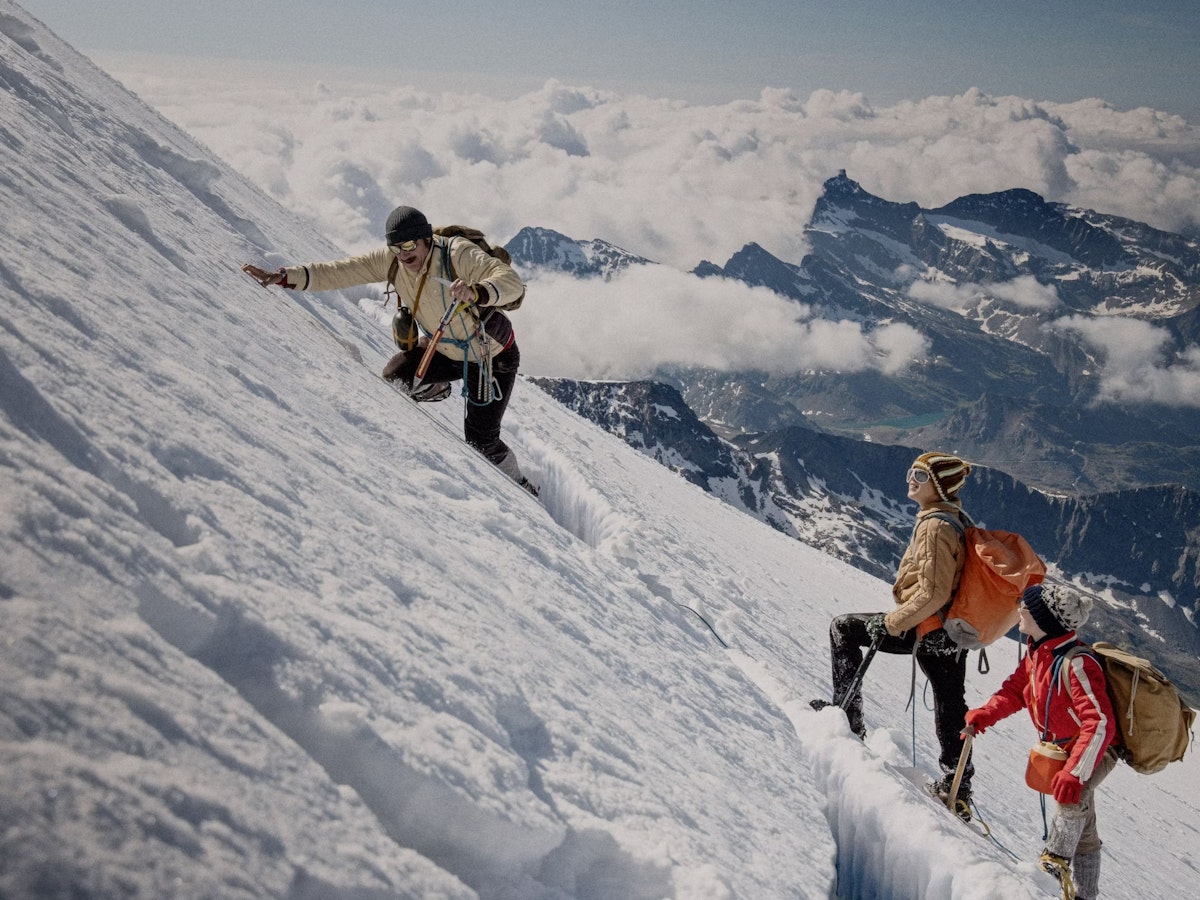 Three climbers with backpacks and winter gear ascend a steep, snowy mountain slope as clouds and rugged peaks stretch out in the background.