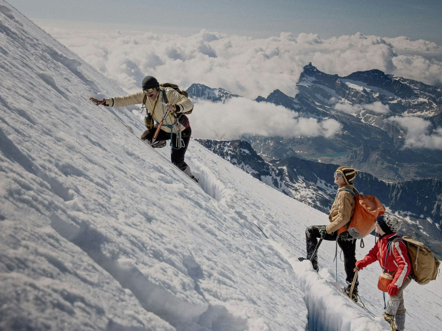 Three climbers with backpacks and winter gear ascend a steep, snowy mountain slope as clouds and rugged peaks stretch out in the background.