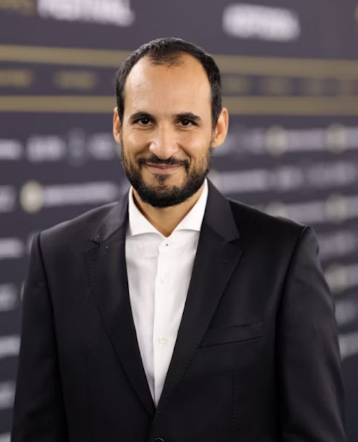 A man with short dark hair and a beard, dressed in a black suit and white shirt, stands smiling in front of a blurred backdrop with horizontal lines and text.