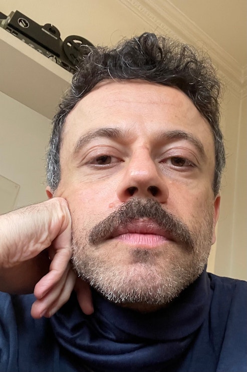 Eine Person mit kurzen lockigen Haaren, Schnurrbart und gestutztem Bart stützt ihr Kinn auf die Hand und blickt direkt in die Kamera. Im Hintergrund sind eine cremefarbene Wand und ein Teil eines Regals zu sehen.