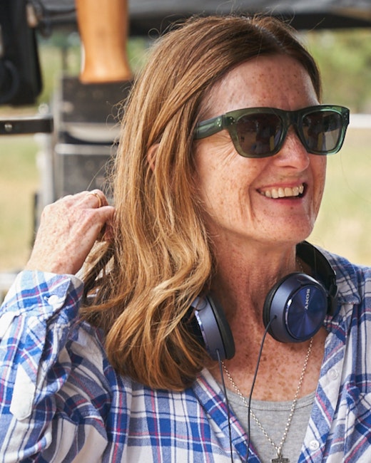 Eine Frau mit Sonnenbrille und blau kariertem Hemd lächelt und berührt ihr Haar. Sie trägt Kopfhörer um den Hals und steht im Freien. Im Hintergrund sind einige Geräte zu sehen.