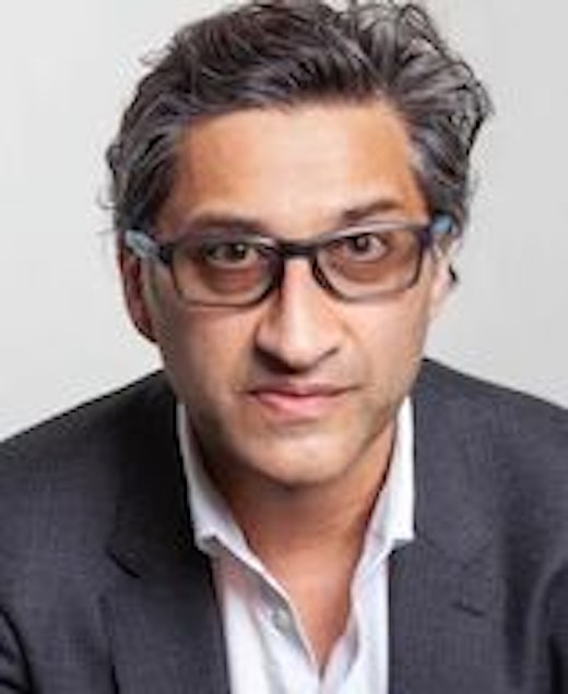 Ein Mann mit kurzem, welligem grauem Haar und Brille, der ein dunkles Jackett über einem weißen Hemd trägt und vor einem schlichten hellen Hintergrund direkt in die Kamera blickt.