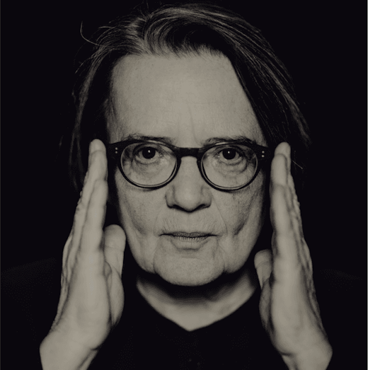 Eine Person mit Brille und schulterlangem Haar blickt in die Kamera und hält beide Hände neben dem Gesicht. Der Hintergrund ist schwarz, das Bild ist schwarz-weiß.