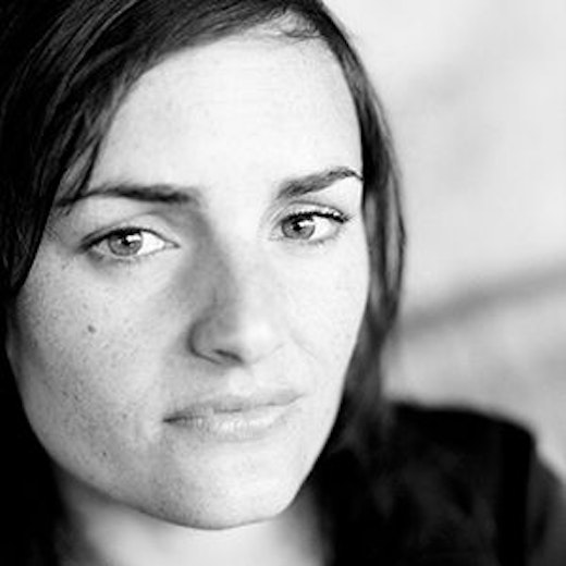 Ein Schwarz-Weiß-Nahaufnahmeporträt einer Person mit mittellangem, dunklem Haar, die mit neutralem Gesichtsausdruck leicht vom Bild abweicht. Der Hintergrund ist unscharf.