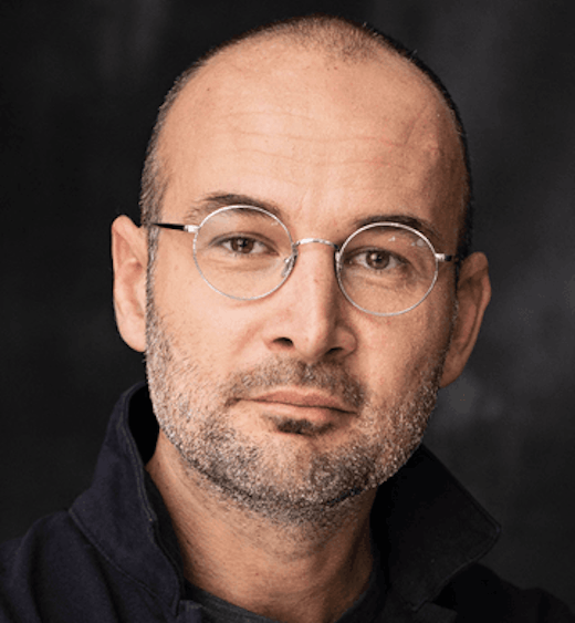 Ein Mann mit rasiertem Kopf, runder Brille und kurzem Bart blickt in die Kamera. Er trägt eine dunkle Jacke und posiert vor einem dunklen, unscharfen Hintergrund.