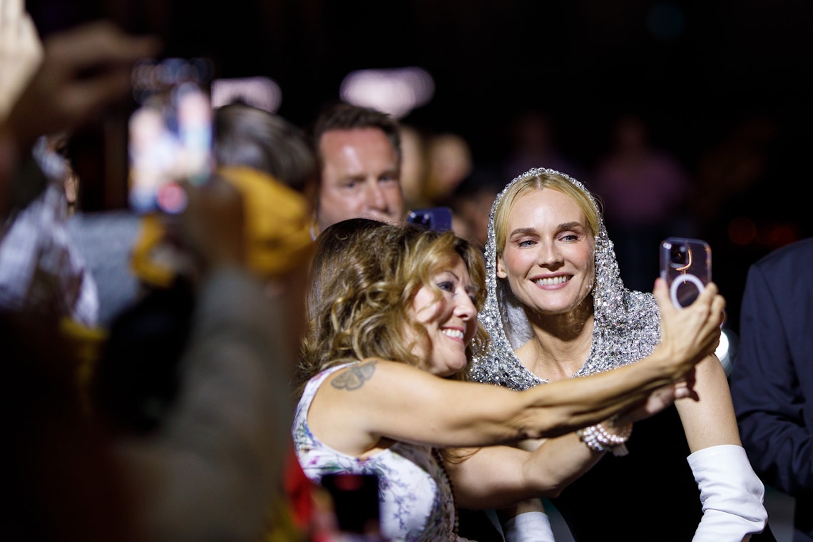 A woman in a sparkling hooded dress poses and smiles for a selfie with a fan in a floral top at a crowded event, surrounded by people taking photos with their phones.