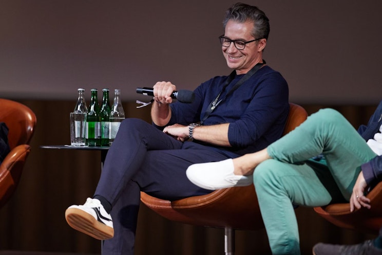 Ein Mann sitzt auf einem braunen Stuhl, hält ein Mikrofon in der Hand und lächelt während einer Podiumsdiskussion. Er trägt eine Brille, ein dunkles Hemd und Turnschuhe. Wasserflaschen stehen auf einem kleinen Tisch neben ihm. Eine weitere Person sitzt in der Nähe, teilweise sichtbar.