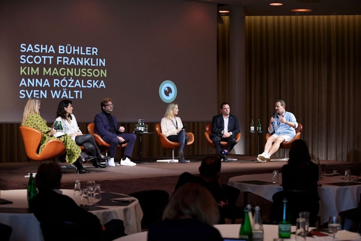 Sechs Personen sitzen mit Mikrofonen und Wasserflaschen auf der Bühne bei einer Podiumsdiskussion. Dahinter sind auf einem Bildschirm fünf Namen in Fettdruck zu sehen: Sasha Bühler, Scott Franklin, Kim Magnusson, Anna Różalska und Sven Wälti. Das Publikum schaut zu.