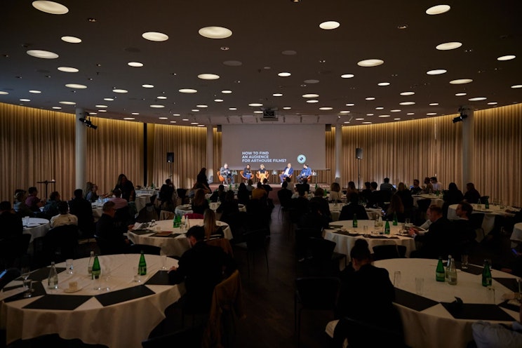 Ein Konferenzraum mit runden Tischen und Teilnehmern, die einer Podiumsdiskussion auf der Bühne lauschen. Auf dem Bildschirm hinter den Diskussionsteilnehmern wird das Thema „Lohnlücken für alle schneller finden und beheben“ angezeigt.