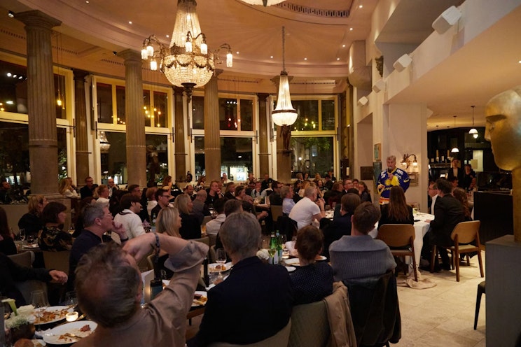 Eine große Gruppe von Menschen sitzt an Tischen in einem eleganten Restaurant mit hohen Decken, Kronleuchtern und großen Fenstern und verfolgt aufmerksam einen Redner vorne im Raum.