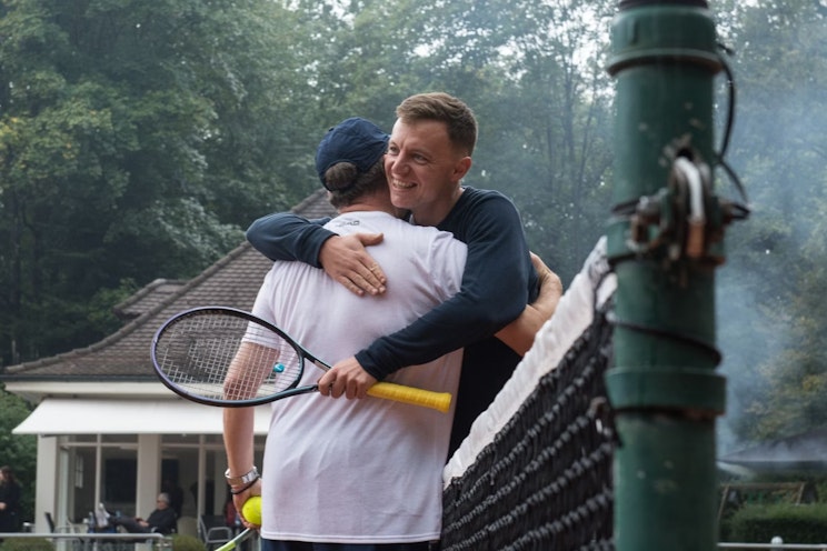 Zwei Personen auf einem Tennisplatz umarmen sich freundschaftlich über das Netz hinweg. Eine Person hält einen Tennisschläger in der Hand. Im Hintergrund sind an einem bewölkten Tag Bäume und ein Gebäude zu sehen.