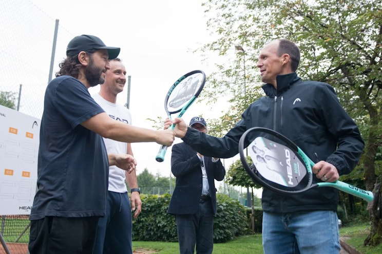 Drei Männer stehen draußen auf einem Tennisplatz, lächeln und schütteln Hände, während sie Tennisschläger in den Händen halten. Eine weitere Person im Hintergrund klatscht. Hinter ihnen sind Bäume und ein Zaun zu sehen.