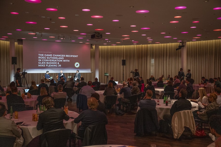 A large audience sits at round tables in a modern event space, watching a panel discussion onstage. The screen behind the speakers reads: The Game Changer Recipient Roe Sutherland in conversation with Glenn Basner & Mike Fleming Jr.