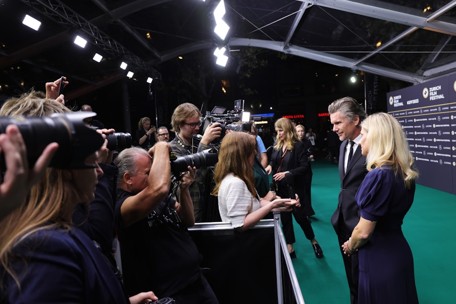 Celebrities pose on a green carpet under a transparent tent as photographers and reporters surround them, taking photos and conducting interviews at a film festival event.