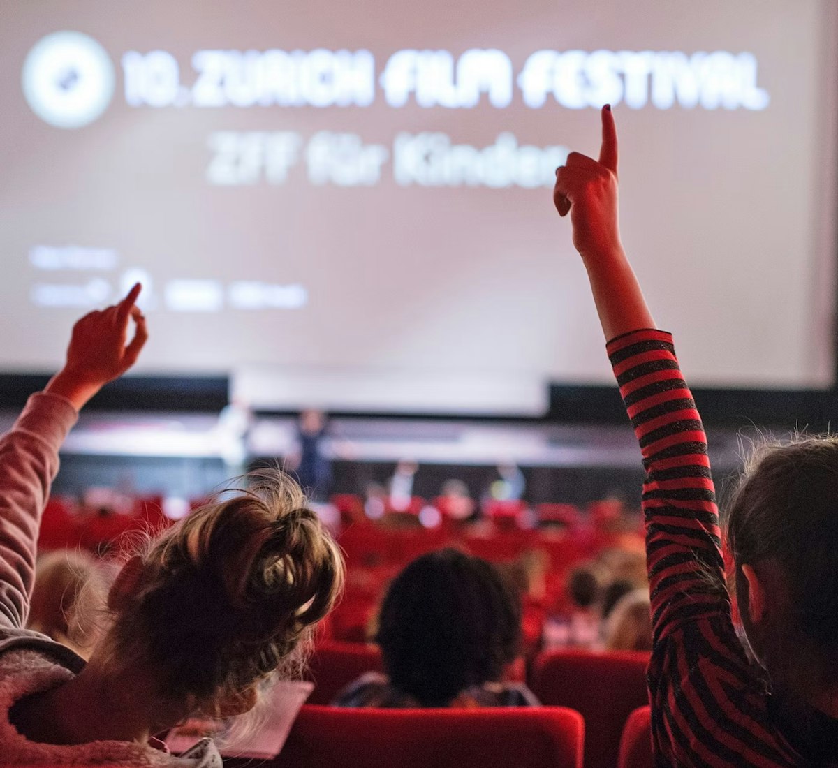 Kinder sitzen in einem Kino mit roten Sitzen und heben ihre Hände in Richtung einer grossen Leinwand, auf der das 10. Zürcher Filmfestival ZFF für Kinder. Die Atmosphäre ist lebendig und einnehmend.