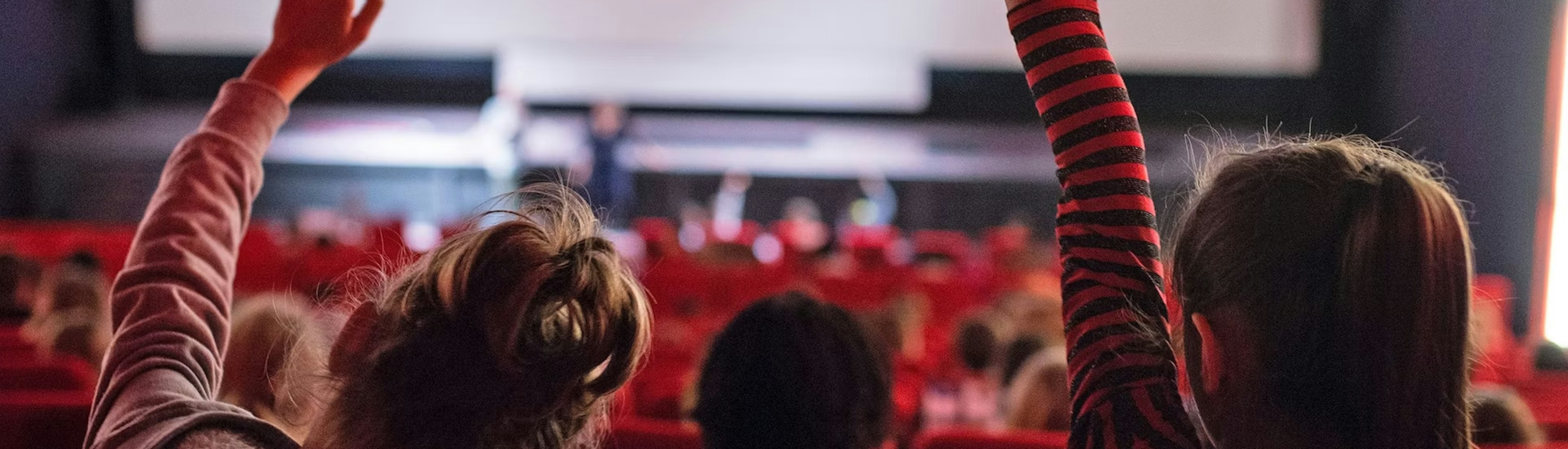Kinder sitzen in einem Kino mit roten Sitzen und heben ihre Hände in Richtung einer grossen Leinwand, auf der das 10. Zürcher Filmfestival ZFF für Kinder. Die Atmosphäre ist lebendig und einnehmend.