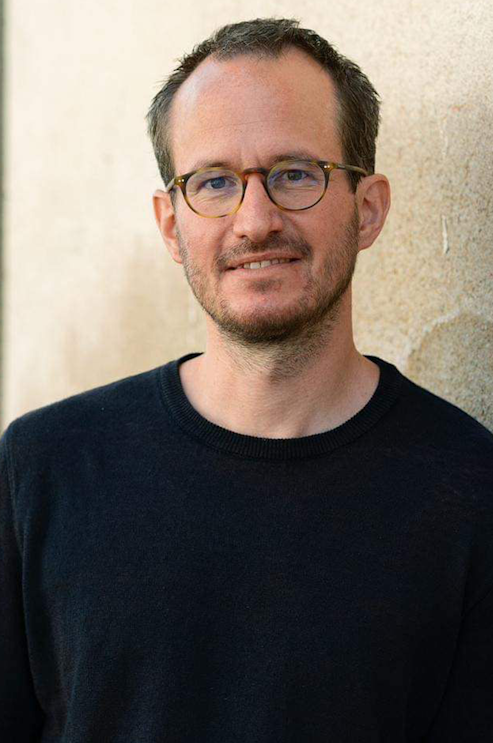 Ein Mann mit kurzen braunen Haaren, Brille und gestutztem Bart trägt einen schwarzen Pullover, steht vor einer hellen, strukturierten Wand und lächelt sanft in die Kamera.