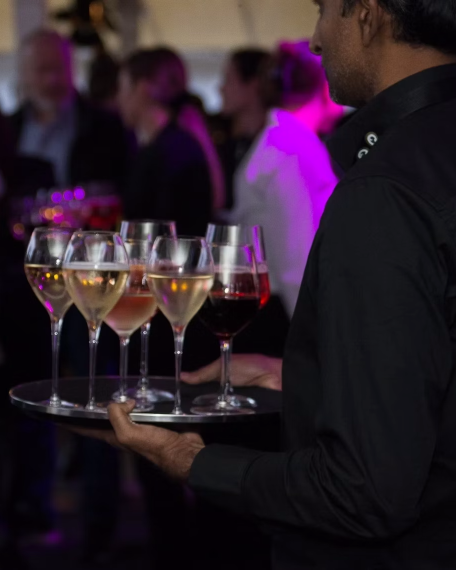 Ein Kellner in einem dunklen Hemd trägt bei einer gut besuchten Veranstaltung ein Tablett mit mehreren Gläsern Wein und Champagner, im unscharfen Hintergrund mischen sich die Leute.