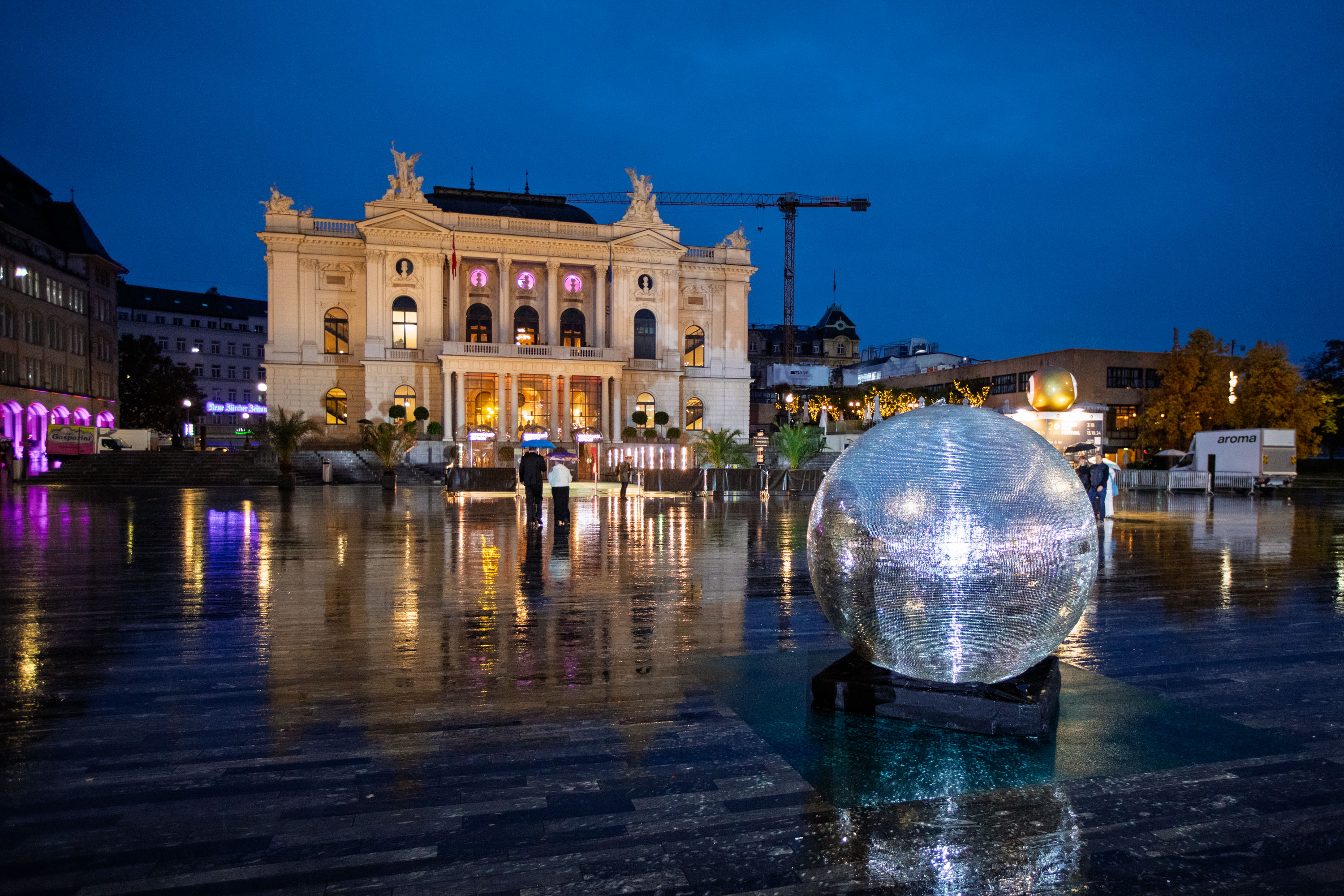 Eine große reflektierende Kugel steht nachts auf einem nassen Platz, mit einem großen beleuchteten Gebäude im Hintergrund und den Lichtern der Stadt, die sich auf dem Boden spiegeln.