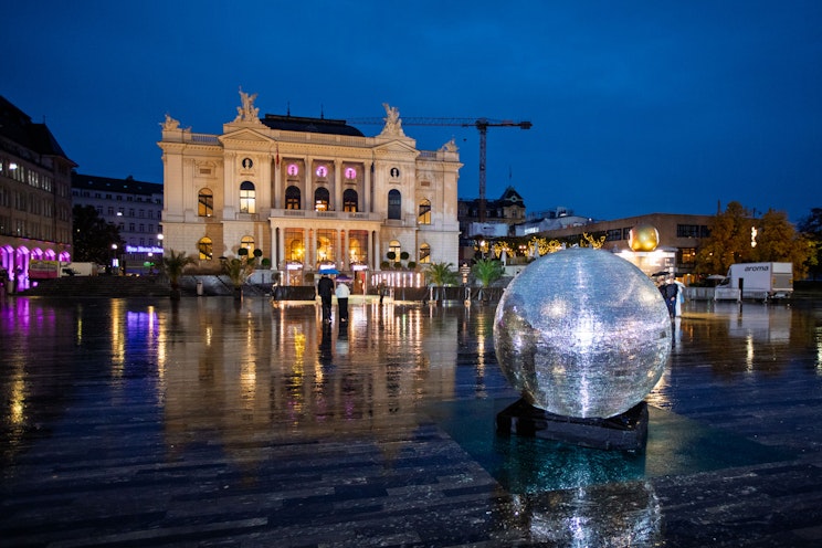 Eine große reflektierende Kugel steht nachts auf einem nassen Platz, mit einem großen beleuchteten Gebäude im Hintergrund und den Lichtern der Stadt, die sich auf dem Boden spiegeln.