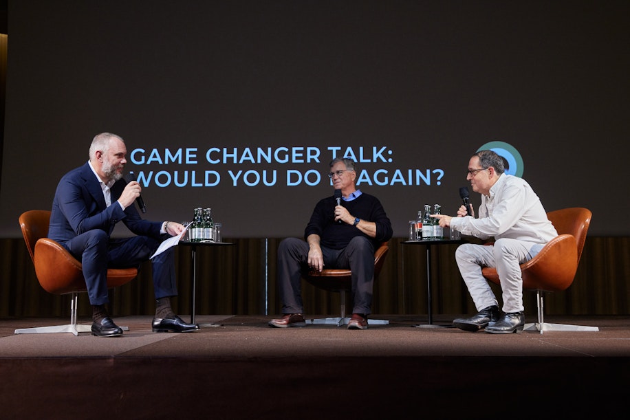 Drei Männer sitzen auf der Bühne und diskutieren an einer Podiumsdiskussion. Einer hält Mikrofon und Notizblock in der Hand, die beiden anderen hören zu. Auf dem Bildschirm hinter ihnen steht: GAME CHANGER TALK: WÜRDEN SIE ES NOCHMAL TUN?.