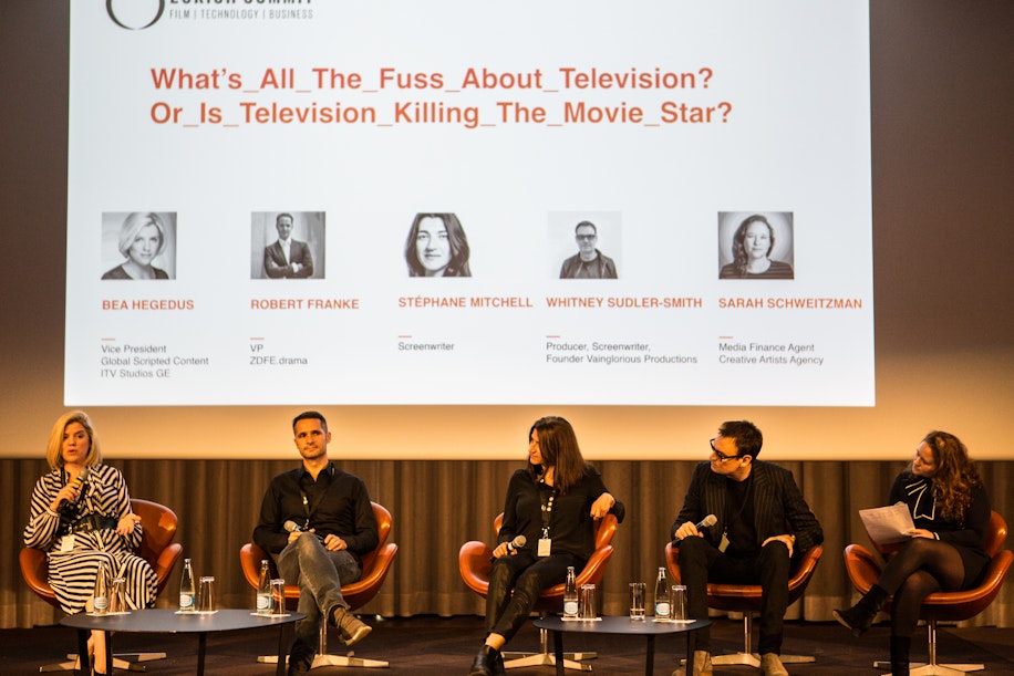 Fünf Diskussionsteilnehmer sitzen mit Mikrofonen auf der Bühne und diskutieren. Hinter ihnen ist eine große Leinwand zu sehen, auf der das Thema „Was ist der ganze Wirbel ums Fernsehen? Oder tötet das Fernsehen den Filmstar?“ sowie ihre Namen und Titel angezeigt werden.