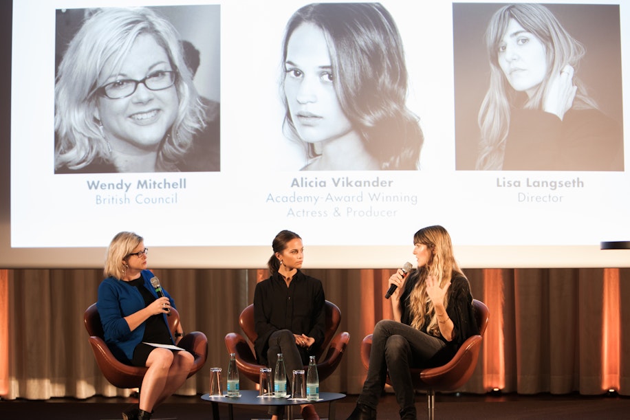 Drei Frauen sitzen auf der Bühne und diskutieren an einer Podiumsdiskussion. Jede hält ein Mikrofon in der Hand. Hinter ihnen werden Bilder und Namen projiziert, die sie als Wendy Mitchell, Alicia Vikander und Lisa Langseth identifizieren.