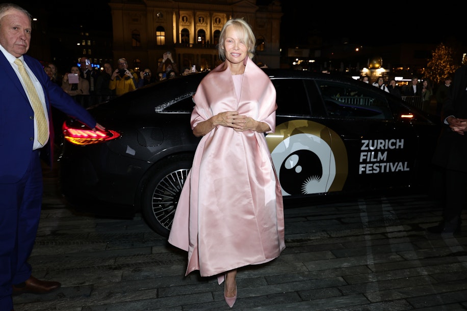 Eine Frau in einem blassrosa Kleid und Schal posiert nachts vor einem schwarzen Auto mit dem Logo des „Zurich Film Festival“, während ein Mann in einem blauen Anzug daneben steht. Im Hintergrund sind helle Lichter der Stadt und Gebäude zu sehen.
