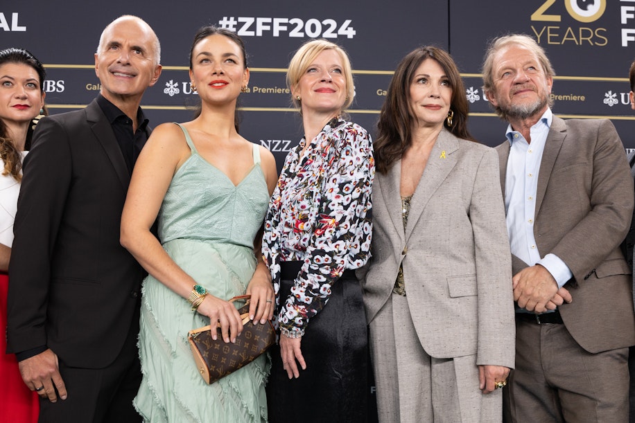 Eine Gruppe von sechs Personen in Abendgarderobe posiert gemeinsam auf einem roten Teppich beim Zurich Film Festival 2024, im Hintergrund sind die Banner und Logos der Veranstaltung zu sehen.
