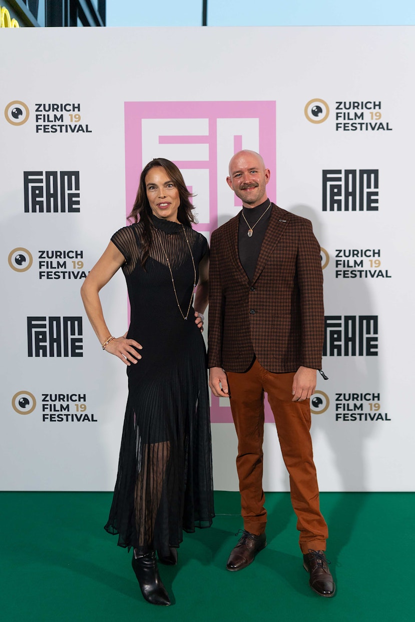 Eine Frau im schwarzen Kleid und ein Mann im braunkarierten Blazer posieren gemeinsam auf dem grünen Teppich des Zurich Film Festival vor einer Markenkulisse.