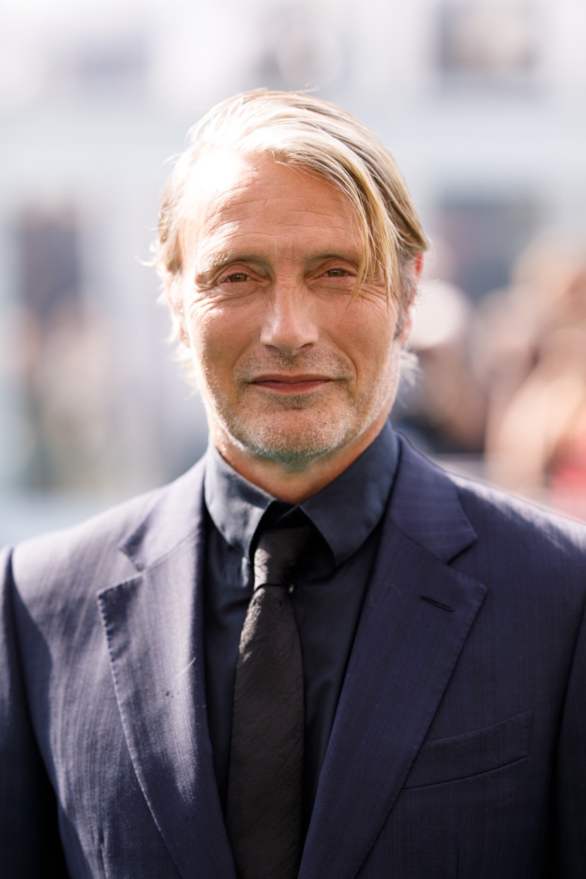 Ein Mann mit hellem Haar und einem leichten Lächeln, der einen dunklen Anzug, ein Hemd und eine Krawatte trägt, steht im Freien vor einem unscharfen Hintergrund aus Menschen und Gebäuden.