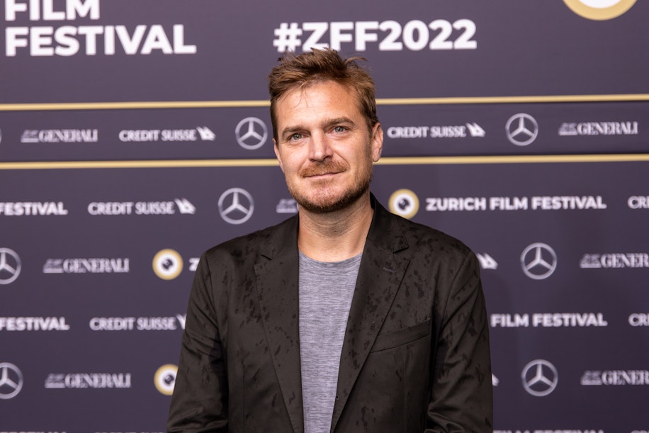 Ein Mann im dunklen Blazer steht vor einer Kulisse mit Logos und den Aufschriften ZURICH FILM FESTIVAL und #ZFF2022. Er blickt in die Kamera und lächelt leicht.