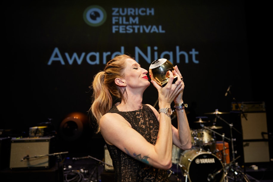 Eine Frau in einem schwarzen Kleid küsst freudig eine goldene Trophäe auf der Bühne, während auf der Leinwand hinter ihr die Award Night des Zurich Film Festivals läuft und im Hintergrund Musikinstrumente zu sehen sind.
