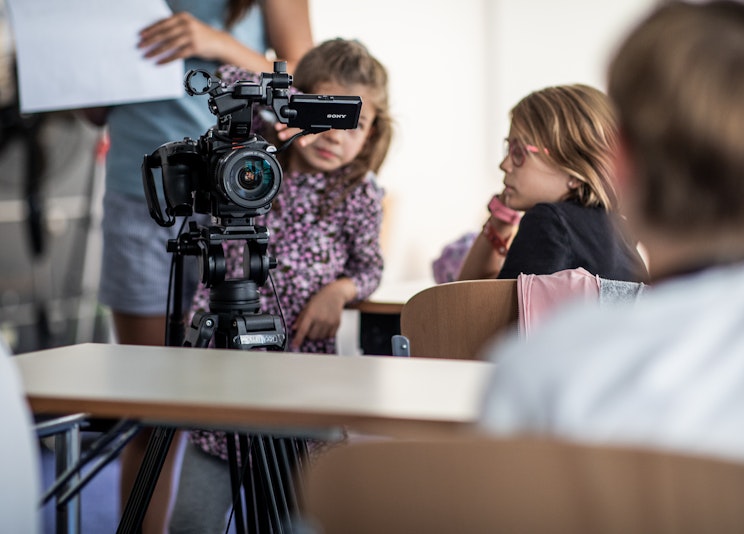 Zwei junge Mädchen stehen in einem Klassenzimmer hinter einer Videokamera auf einem Stativ. Ein Mädchen trägt eine Brille und sitzt an einem Schreibtisch, während das andere neben der Kamera steht und sie interessiert betrachtet.