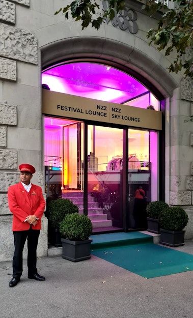 Ein Türsteher in roter Uniform und Mütze steht vor dem Eingang eines großen Gebäudes mit der Aufschrift „NZZ Festival Lounge“ und „NZZ Sky Lounge“. Es hat Glastüren, violette Beleuchtung im Inneren und Topfpflanzen neben der Tür.