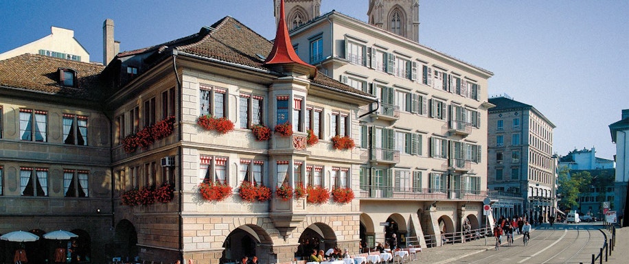 Historische europäische Gebäude mit weißen Fassaden und grünen Fensterläden sowie roten Blumen in Blumenkästen. Menschen sitzen an Tischen im Freien unter Bögen entlang einer Kopfsteinpflasterstraße. Im Hintergrund ist ein Kirchturm zu sehen.