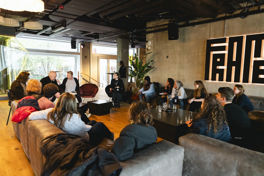 In einer modernen Lounge sitzt eine Gruppe von Menschen kreisförmig auf Sofas und Stühlen und unterhält sich. Große Fenster und eine moderne Einrichtung schaffen eine entspannte, offene Atmosphäre.